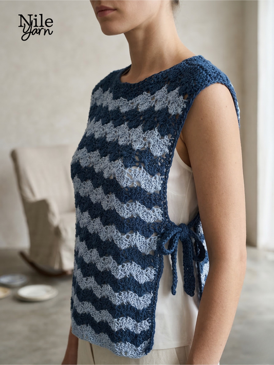 A woman wearing a crocheted "Sea Breeze" side-tie top made from blue and light blue wave-patterned Egyptian cotton yarn. The top features a sleeveless, open-side design with a functional bow tie at the waist.