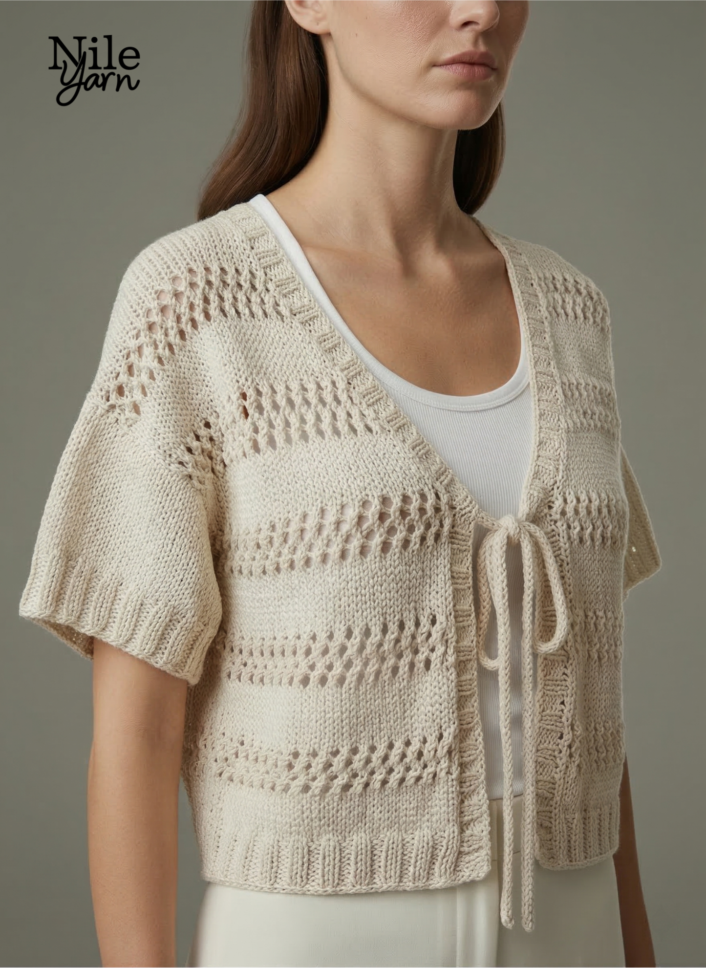 A woman wearing a cream-colored, short-sleeved knitted vest with horizontal eyelet lace patterns and a front tie closure over a white top.