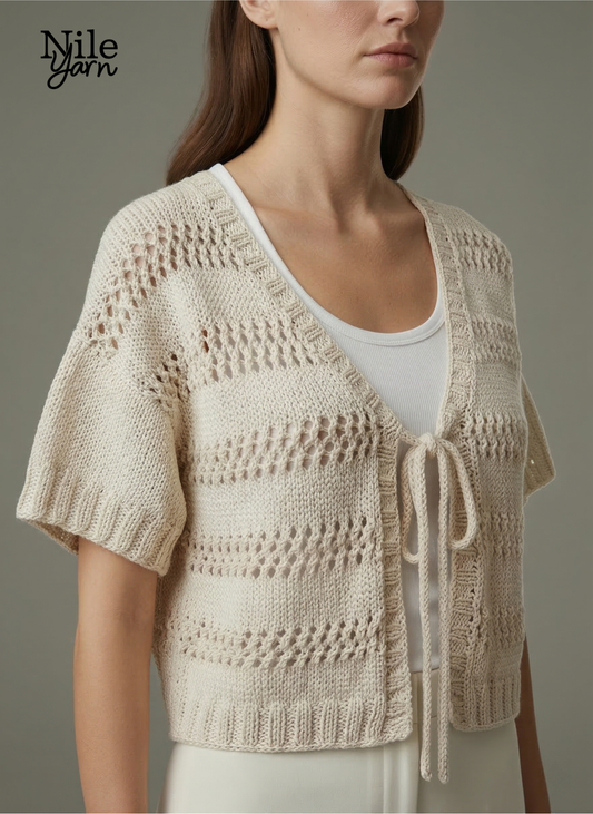 A woman wearing a cream-colored, short-sleeved knitted vest with horizontal eyelet lace patterns and a front tie closure over a white top.