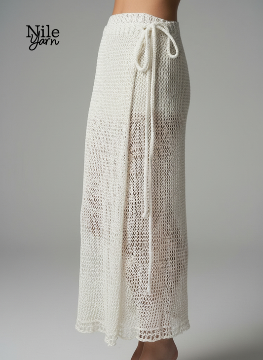 Side view of a woman wearing a long, white, hand-knitted wrap skirt with a decorative tie at the waist and a semi-sheer, open-knit texture.